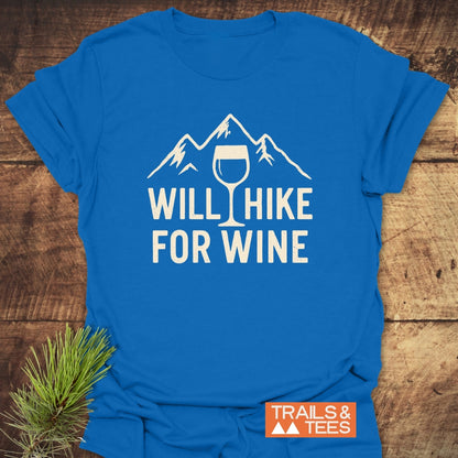 Will Hike For Wine T-Shirt with mountain and wine glass design, laid on wood surface. Features soft ring-spun cotton, durable print, and Trails & Tees logo, embodying outdoor adventure spirit.