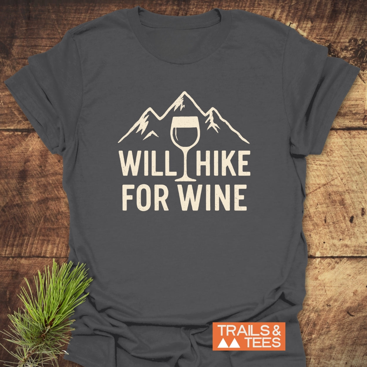 Will Hike For Wine T-Shirt with a mountain and wine glass design printed on dark-gray fabric, showcasing a nature-inspired slogan, laid flat with a pine bough accentuating the outdoor theme.