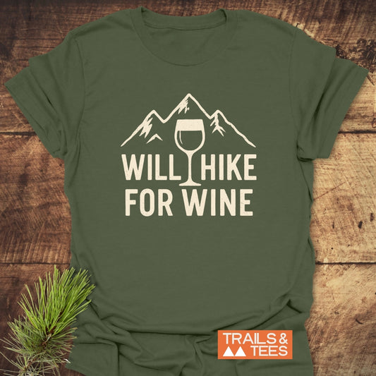 Will Hike For Wine T-Shirt featuring a stylized mountain and wine glass design in off-white, laid flat. Includes a small orange TRAILS & TEES logo and a pine sprig accent.