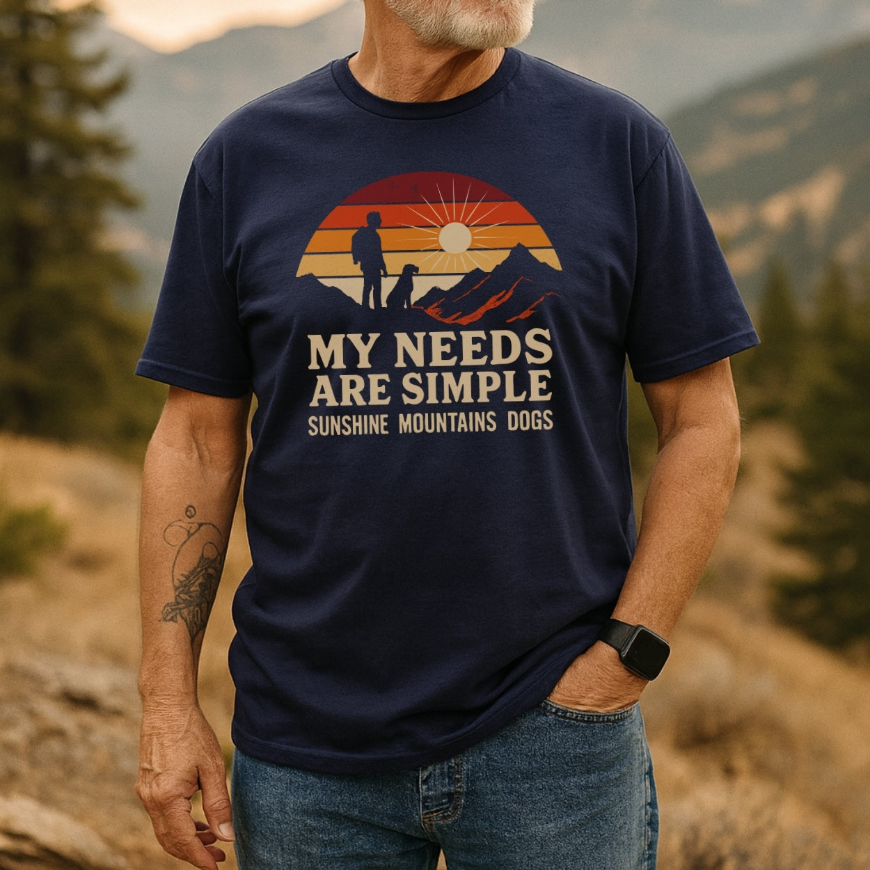 Man in mountain setting wears the Simple Needs Vintage T-Shirt featuring a retro graphic of a person and dog on a ridge with sunset stripes, embodying outdoor adventure and comfort.