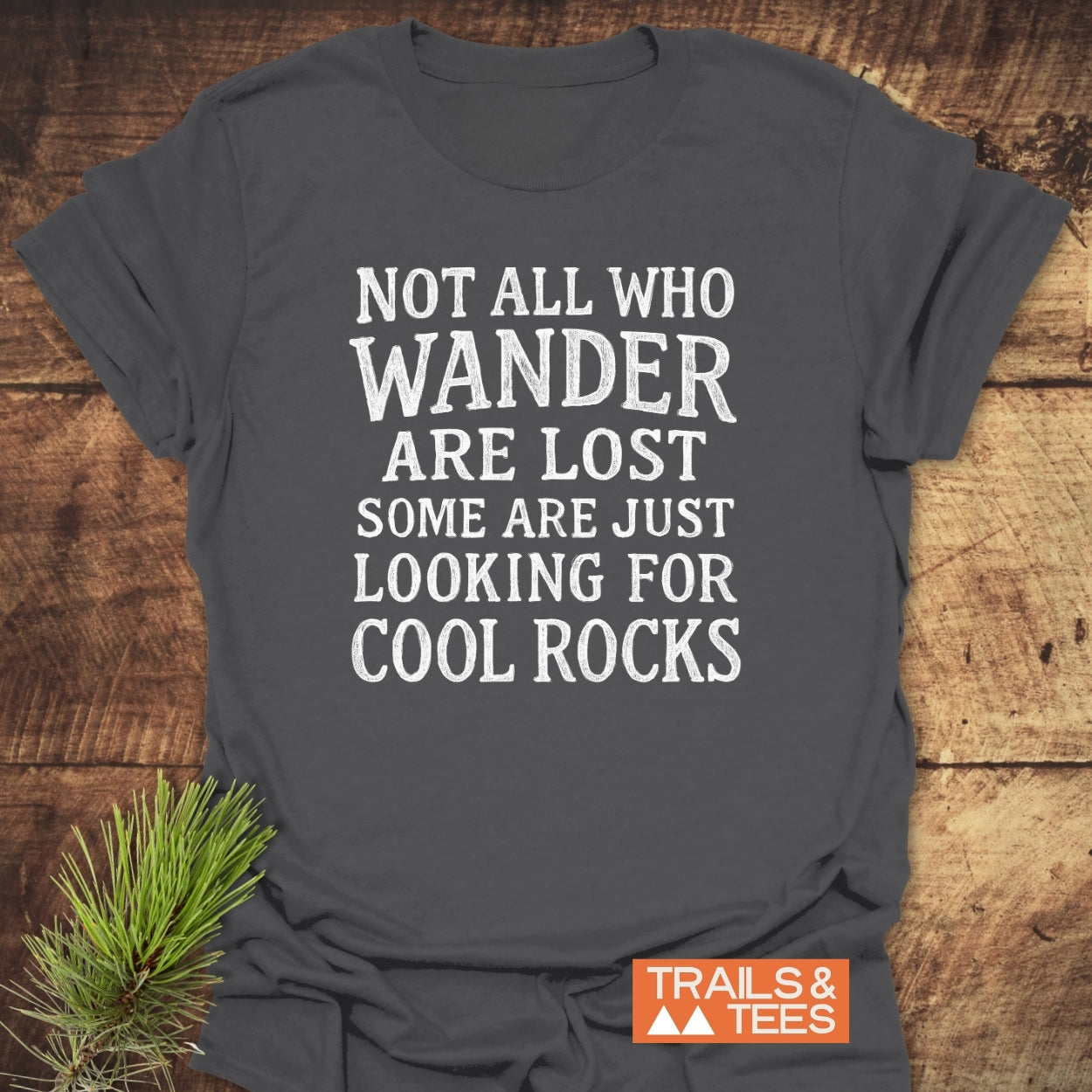 Not All... Rustic Chalkboard T-Shirt on a wooden surface, featuring a white slogan about wandering and cool rocks. Includes the Trails & Tees logo, emphasizing outdoor-inspired comfort and durability.