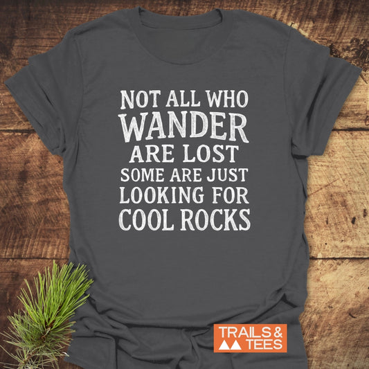 Not All... Rustic Chalkboard T-Shirt on a wooden surface, featuring a white slogan about wandering and cool rocks. Includes the Trails & Tees logo, emphasizing outdoor-inspired comfort and durability.
