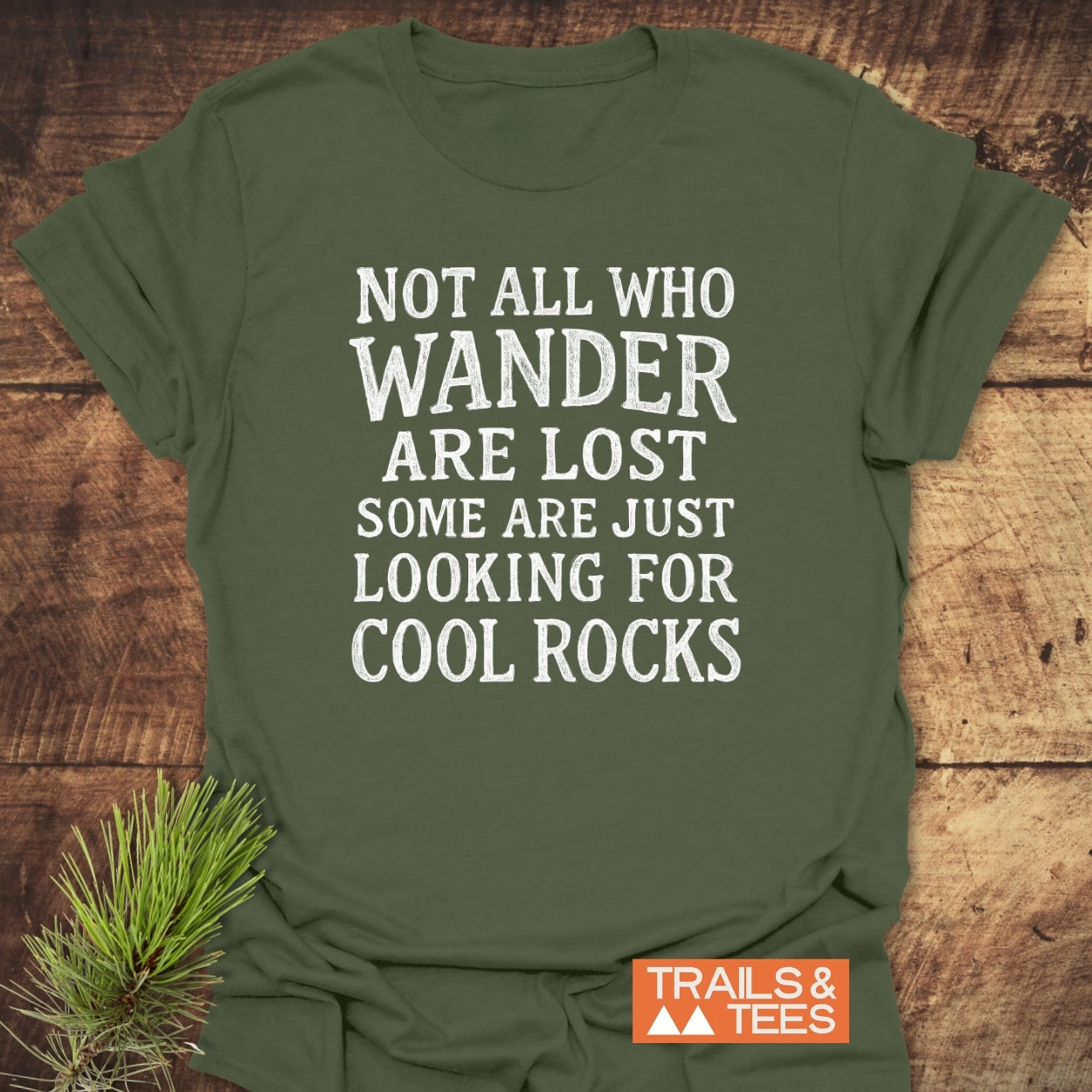 Not All Who Wander Are Lost Rustic Chalkboard T-Shirt, featuring white distressed text on olive-green fabric, complemented by an orange TRAILS & TEES. logo, showcased on a wooden surface.