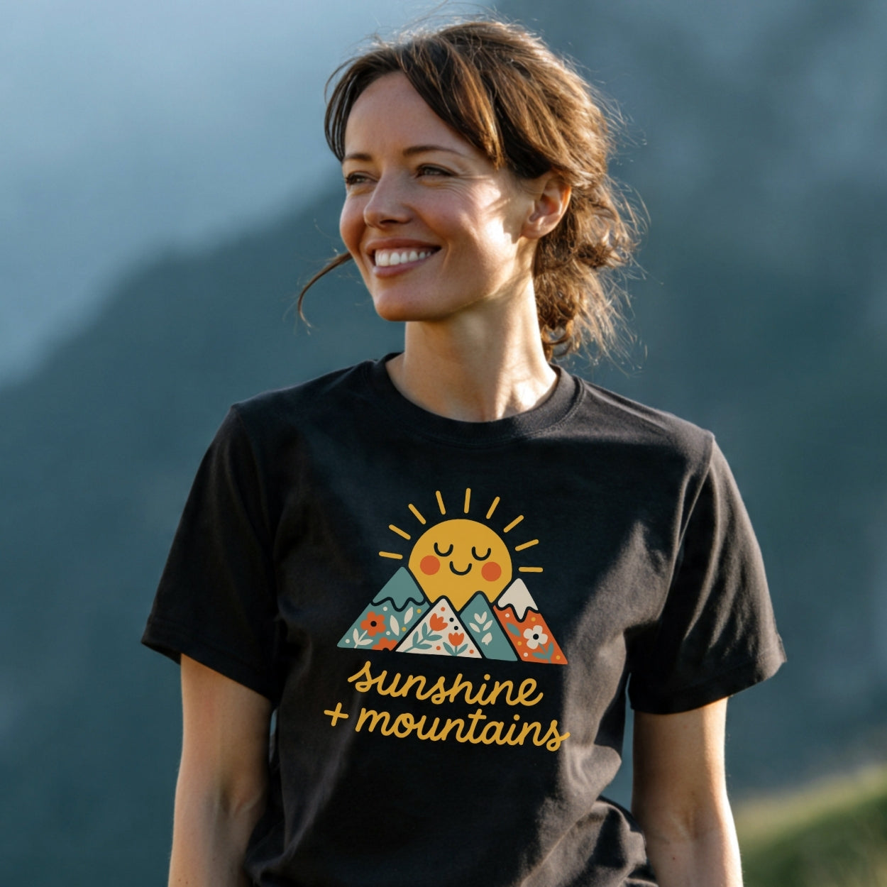 Young woman wearing the Sunshine And Mountains T-Shirt, featuring a cheerful sun rising behind floral-patterned peaks. The black tee highlights outdoor adventures with soft, lightweight cotton for comfort and durability.