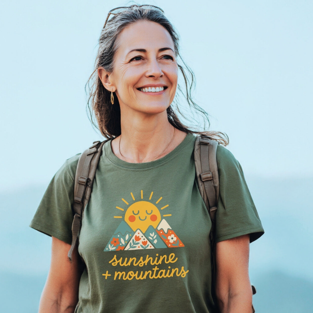 Woman outdoors in a khaki-green Sunshine And Mountains T-Shirt, featuring a smiling sun and patterned mountains graphic, paired with a daypack. Perfect for everyday adventures and outdoor enthusiasts.