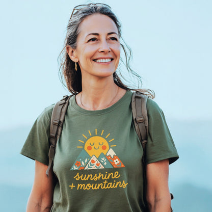 Woman outdoors in a khaki-green Sunshine And Mountains T-Shirt, featuring a smiling sun and patterned mountains graphic, paired with a daypack. Perfect for everyday adventures and outdoor enthusiasts.