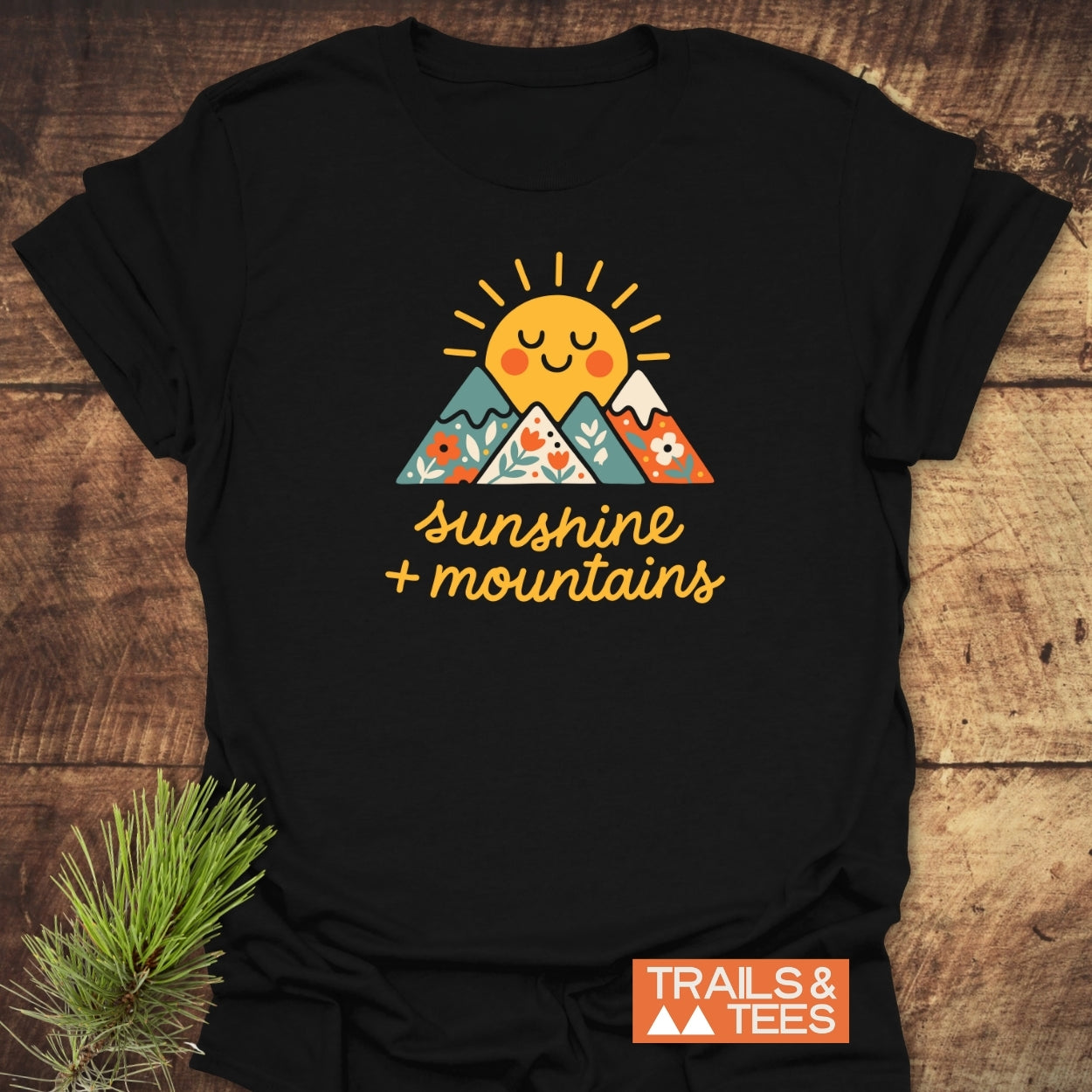 Sunshine And Mountains T-Shirt featuring a cheerful yellow sun and flower-patterned mountains graphic, laid flat on wood with a pine sprig beside it, showcasing Trails & Tees' adventurous spirit.