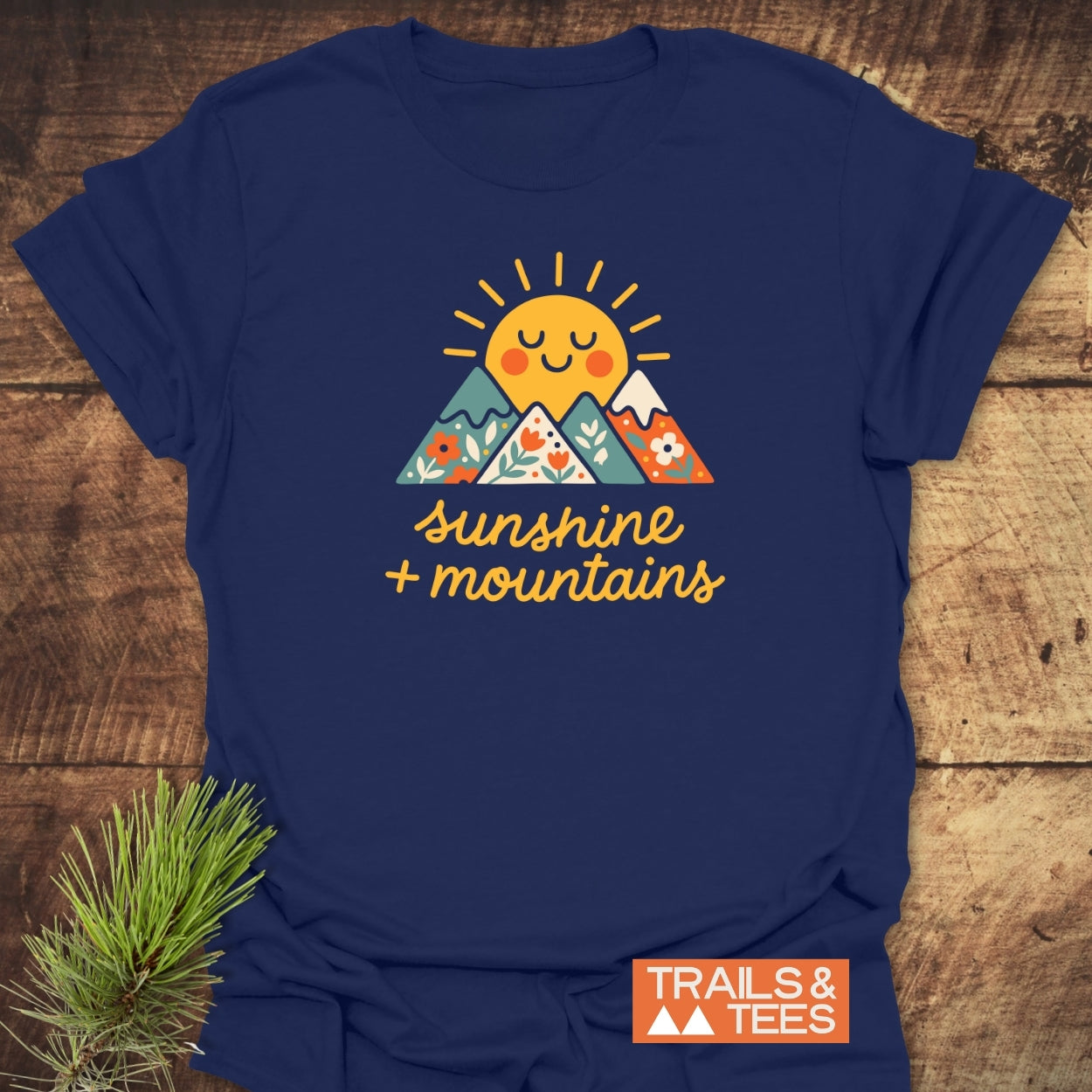 Sunshine And Mountains T-Shirt featuring a cheerful sun over flower-patterned mountains with the slogan “sunshine + mountains.” Includes a small Trails & Tees logo in the corner.