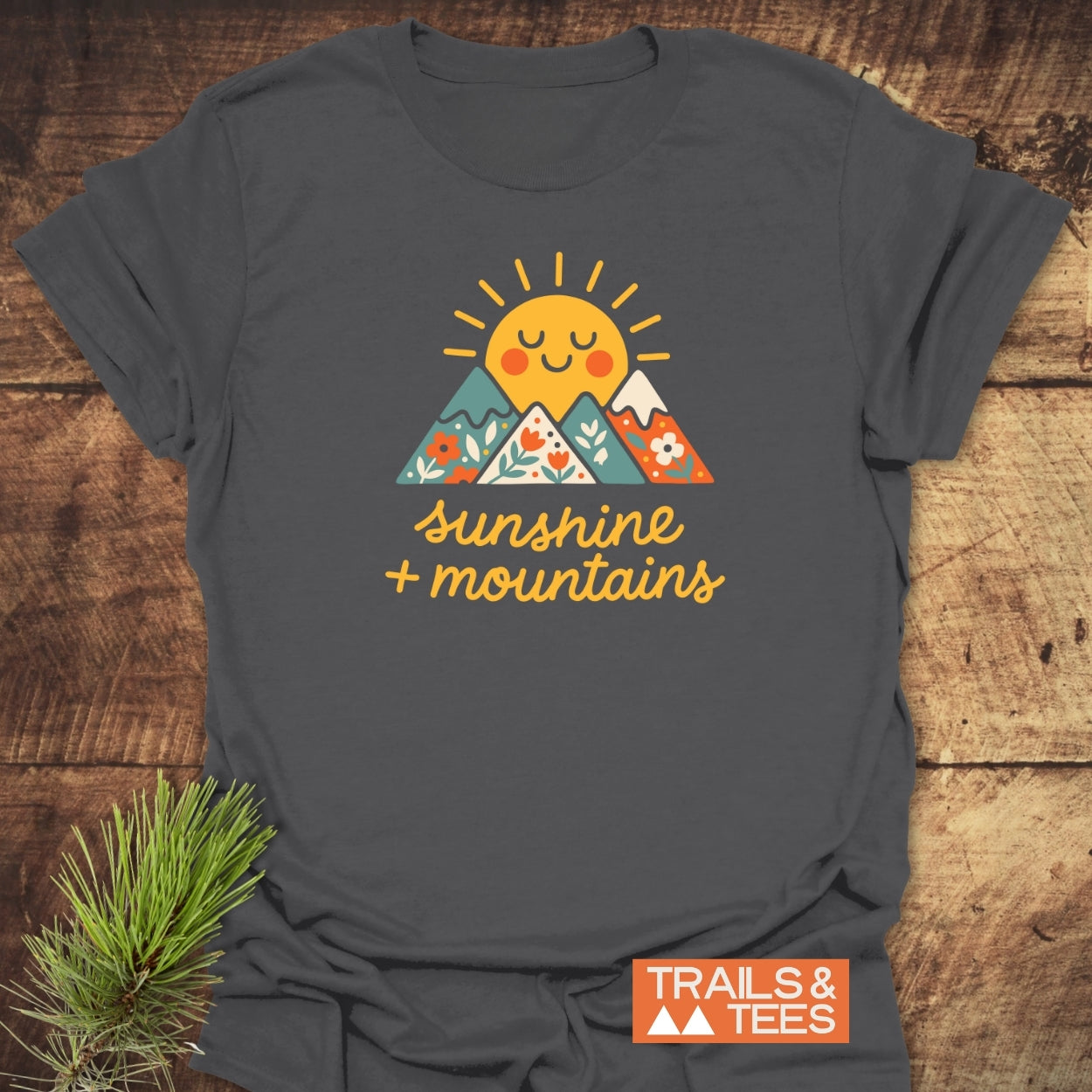 Sunshine And Mountains T-Shirt featuring a graphic of floral-patterned mountains and a cheerful sun. Laid flat on wood, includes TRAILS & TEES logo and a pine sprig for decoration.