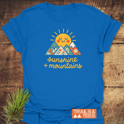Sunshine And Mountains T-Shirt featuring a smiling sun graphic over mountains with floral patterns, hand-lettered text, sunshine + mountains, and a TRAILS & TEES logo on a wooden surface.