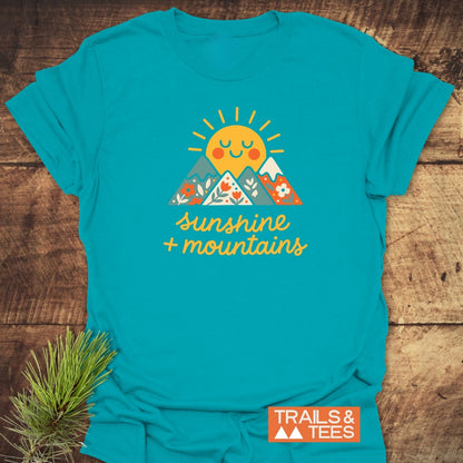 Sunshine And Mountains T-Shirt featuring a cheerful sun and mountain graphic with sunshine + mountains text, on a turquoise shirt. Includes TRAILS & TEES label, emphasizing outdoor adventure and comfort.