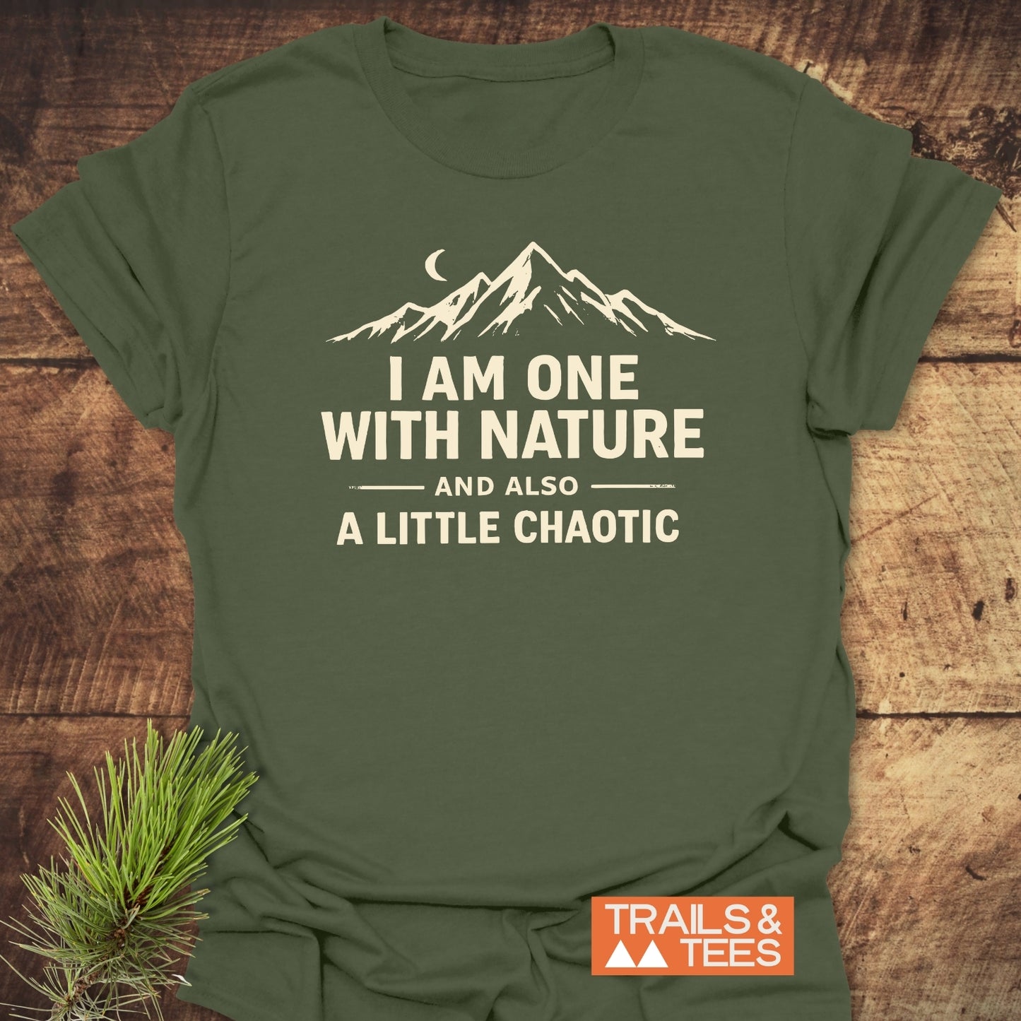 One With Nature T-Shirt featuring a mountain range and crescent moon design, laid on wood with a pine sprig; includes TRAILS & TEES logo, emphasizing adventure-ready comfort and durability.