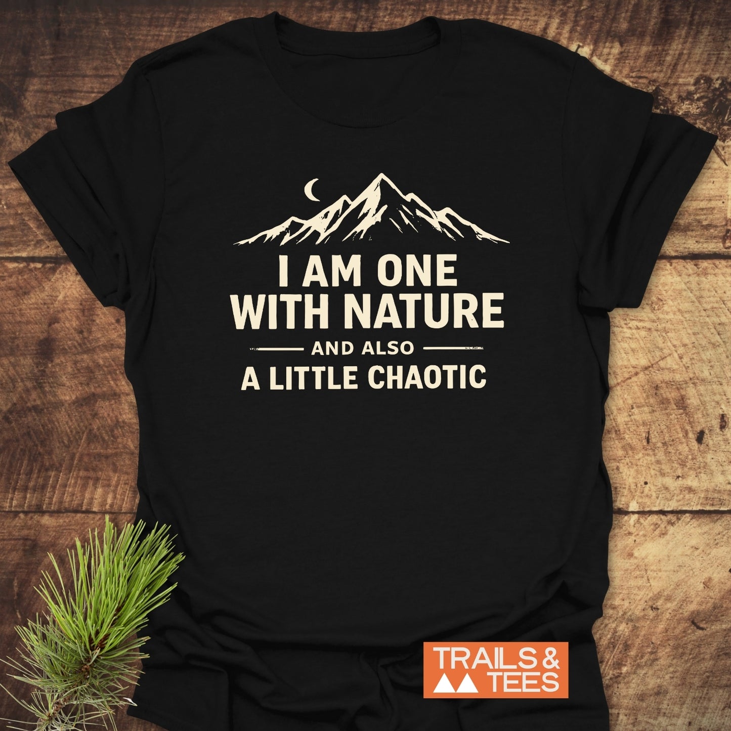 One With Nature T-Shirt, featuring a mountain silhouette and slogan, laid on wood. Includes “TRAILS & TEES.” logo. Designed from soft cotton for comfort, perfect for outdoor enthusiasts.