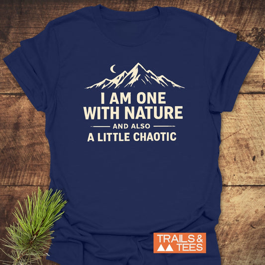 One With Nature T-Shirt featuring a white graphic of a mountain range and crescent moon, with bold text on navy-blue fabric. Includes TRAILS & TEES logo, laid on a wooden surface.