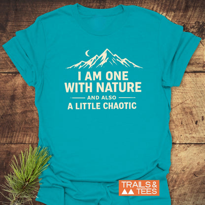 One With Nature T-Shirt featuring a white mountain and crescent moon graphic with a playful slogan, laid on wood. Includes a small orange TRAILS & TEES. label.
