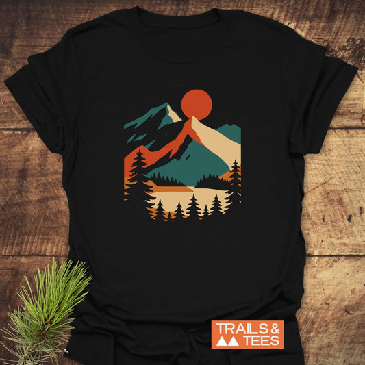 Mountain Sunset Graphic T-Shirt featuring a retro mountain scene with a sun over peaks, lake, and pines on a black shirt, laid flat on wood, with TRAILS & TEES branding nearby.