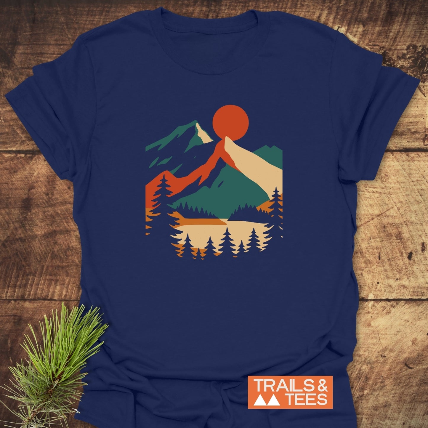 Mountain Sunset Graphic T-Shirt featuring a geometric landscape with pine trees, a lake, mountains, and a red sun. Includes TRAILS & TEES branding and an evergreen sprig beside the shirt.