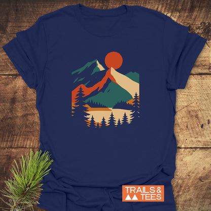 Mountain Sunset Graphic T-Shirt featuring a geometric landscape with pine trees, a lake, mountains, and a red sun. Includes TRAILS & TEES branding and an evergreen sprig beside the shirt.