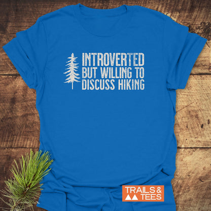 Introverted Hiker T-Shirt featuring INTROVERTED BUT WILLING TO DISCUSS HIKING in distressed white, pine-tree graphic, and Trails & Tees logo, displayed on wood with a pine sprig accent.