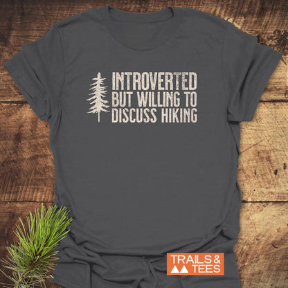 Introverted Hiker T-Shirt with a distressed-white pine tree and hiking message on charcoal-gray fabric, laid flat with a pine sprig and Trails & Tees logo visible.