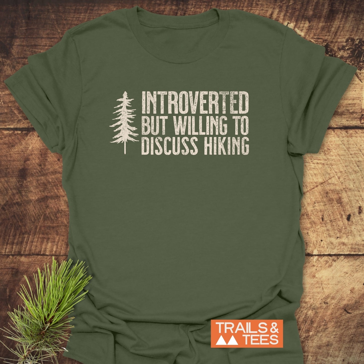 Introverted Hiker T-Shirt featuring a distressed white pine tree print and humorous hiking text, laid on wood. Includes TRAILS & TEES label and pine sprigs for a nature-inspired touch.