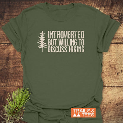 Introverted Hiker T-Shirt featuring a distressed white pine tree print and humorous hiking text, laid on wood. Includes TRAILS & TEES label and pine sprigs for a nature-inspired touch.