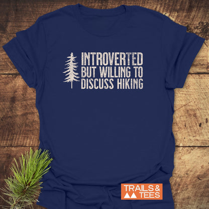 Introverted Hiker T-Shirt featuring a distressed pine tree graphic and text, laid on wood with a pine sprig. Includes TRAILS & TEES logo. Made from soft, pre-shrunk cotton for comfortable adventures.