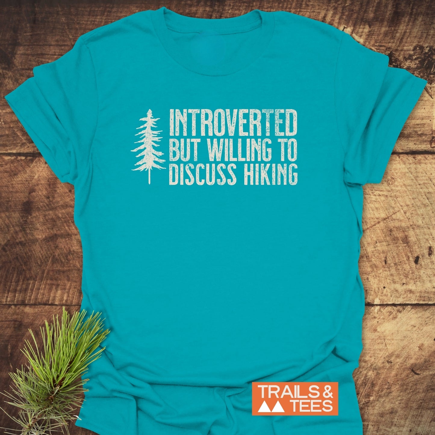Introverted Hiker T-Shirt on wood surface, featuring distressed white text and pine-tree silhouette. Includes orange TRAILS & TEES logo and pine sprig, highlighting outdoor theme and comfort.