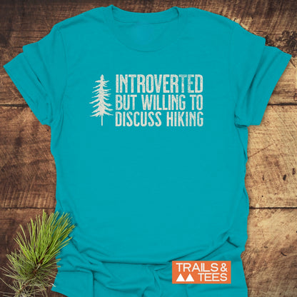 Introverted Hiker T-Shirt on wood surface, featuring distressed white text and pine-tree silhouette. Includes orange TRAILS & TEES logo and pine sprig, highlighting outdoor theme and comfort.
