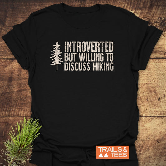 Introverted Hiker T-Shirt featuring a distressed pine tree graphic and text on a soft black fabric, complemented by an orange TRAILS & TEES logo, styled with pine needles for an outdoor theme.