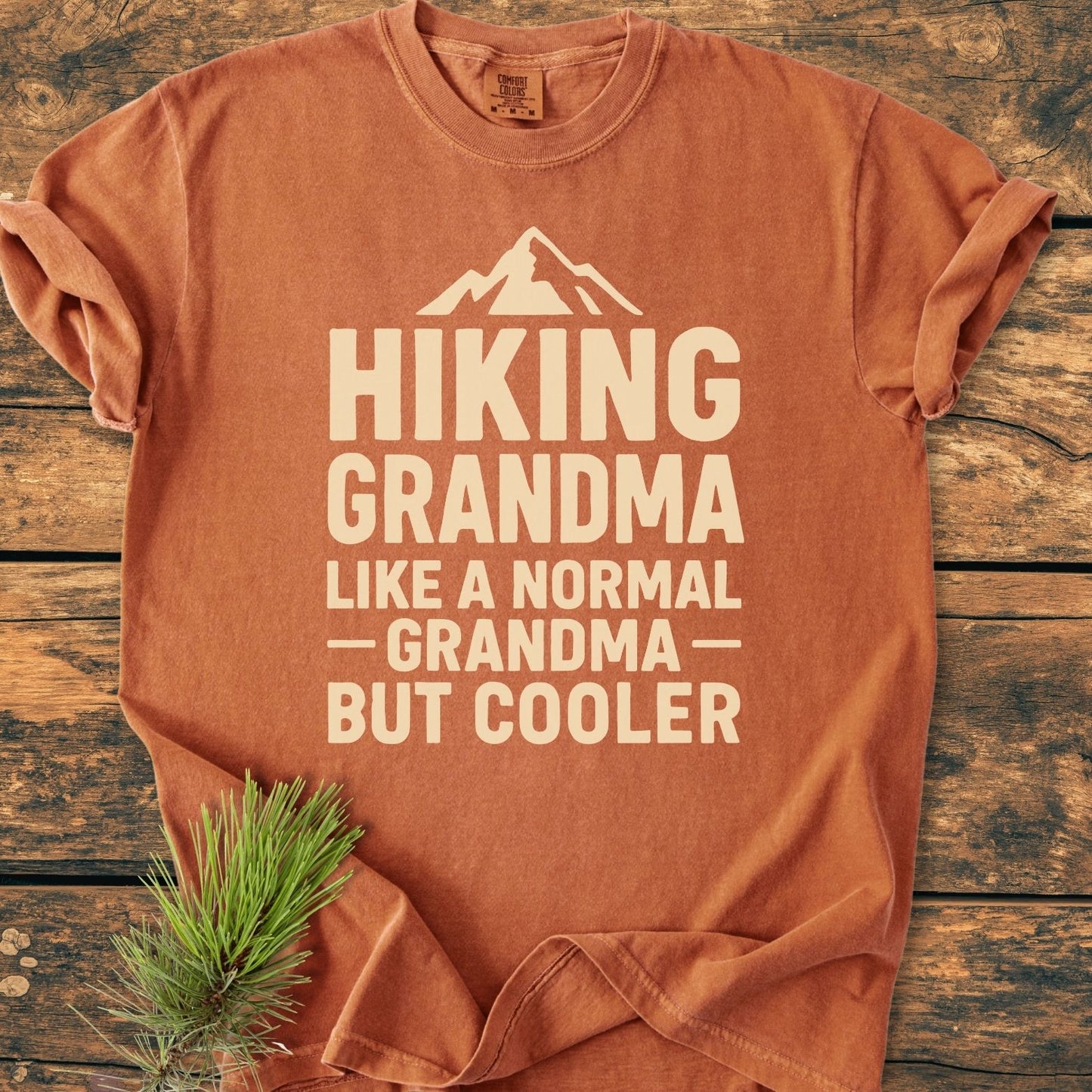 Hiking Grandma Vintage Wash Tee, rust-orange with off-white text and mountain graphic, laid flat on wood. Includes pine sprig for outdoorsy touch. Made from soft, pre-shrunk, 100% ring-spun cotton.