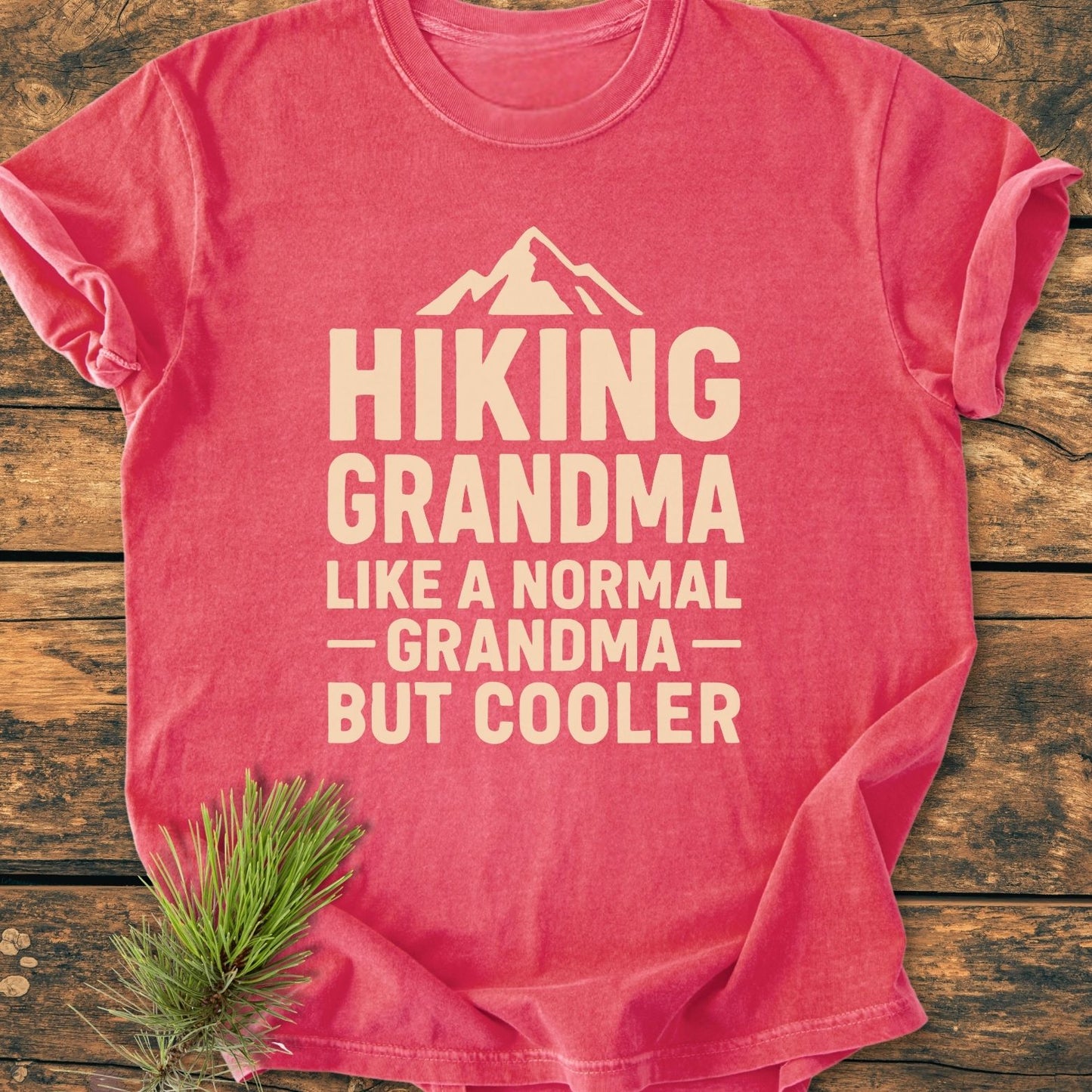 Hiking Grandma Vintage Wash Tee with mountain graphic and playful text on a rustic wooden surface, featuring a soft, relaxed fit from Comfort Colors, accented by a small pine sprig.