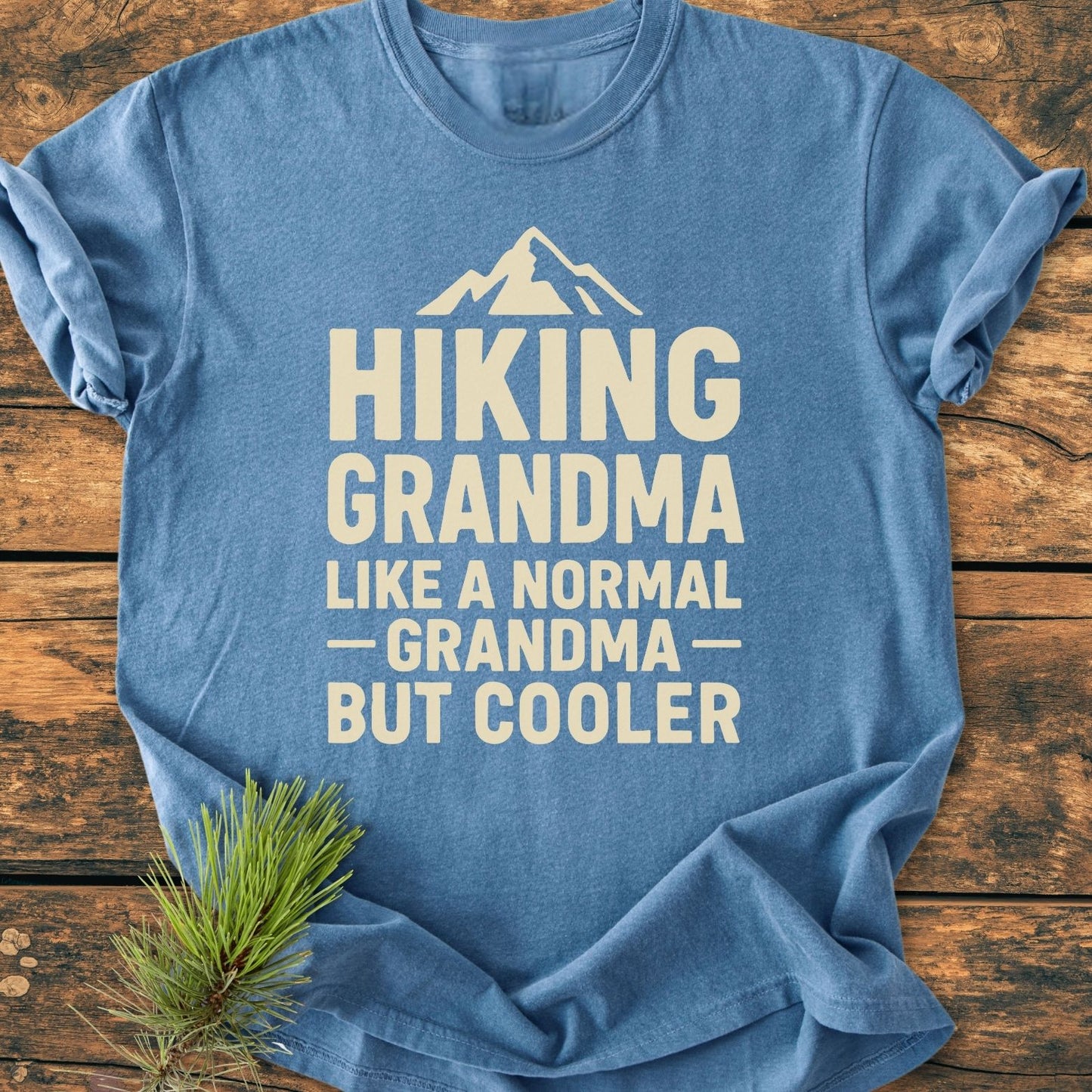 Hiking Grandma Vintage Wash Tee: Heather-blue T-shirt with cream slogan and mountain icon, laid on wood. Features soft, garment-dyed cotton, relaxed fit, and durable stitching. Pine sprig adds an outdoor touch.