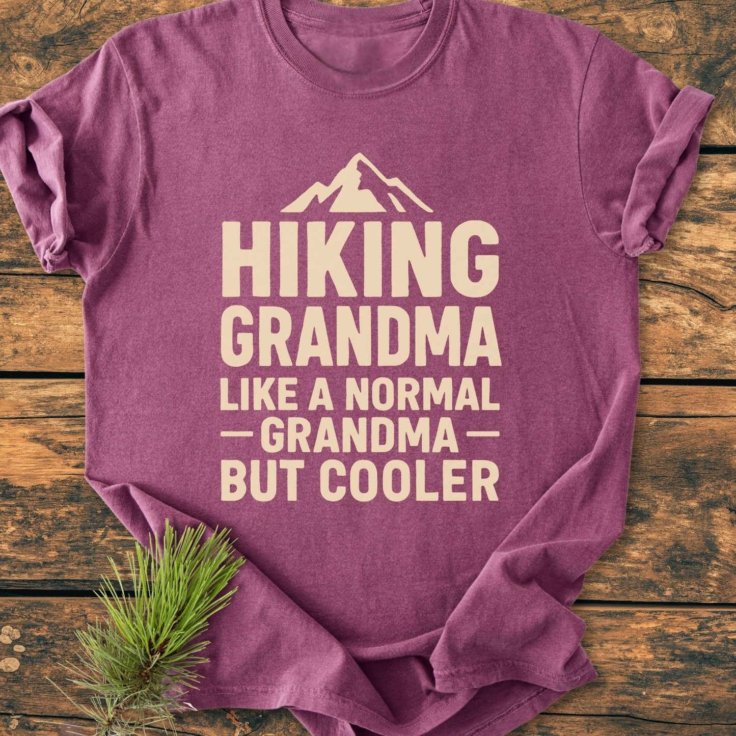 Hiking Grandma Vintage Wash Tee: Purple T-shirt with cream mountain graphic and slogan, 'HIKING GRANDMA, LIKE A NORMAL GRANDMA — BUT COOLER,' styled on wood with pine sprig.