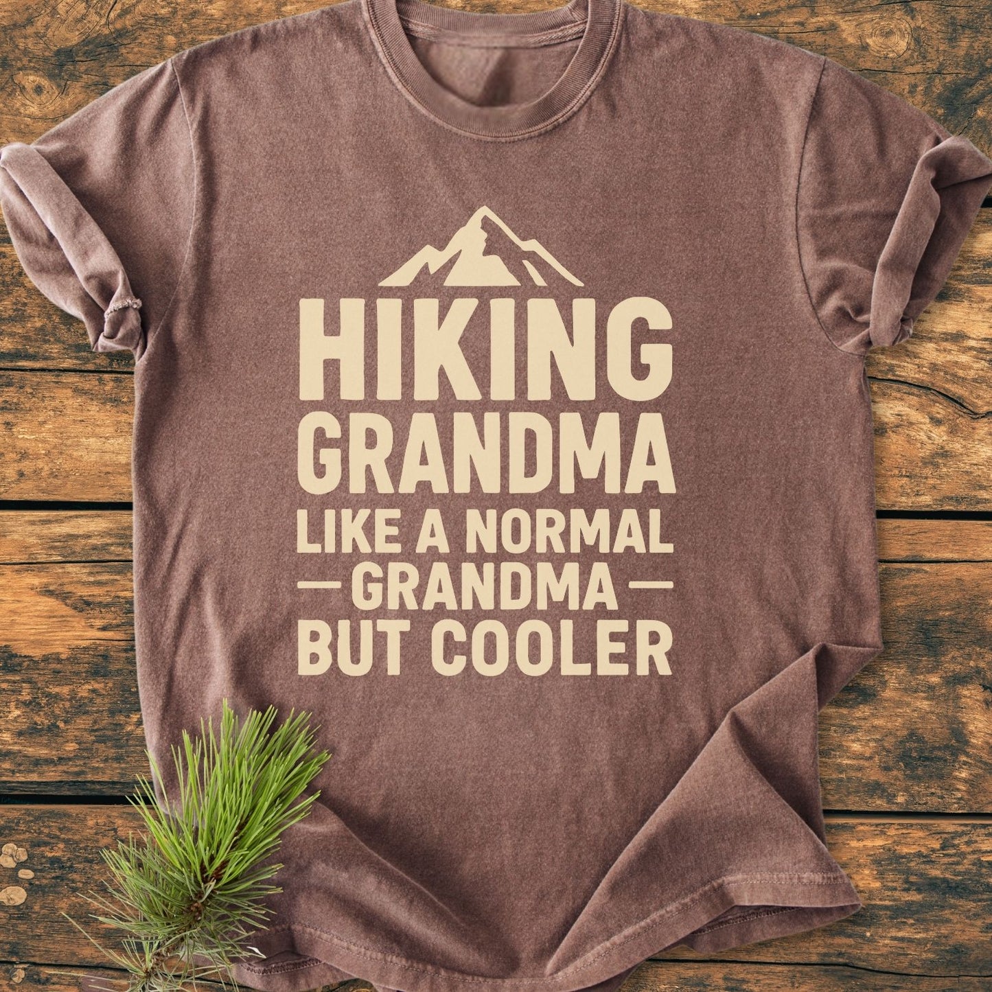Hiking Grandma Vintage Wash Tee displayed on wood, featuring a mountain icon and playful slogan in off-white. Accompanied by a pine needle sprig, highlighting its relaxed fit and garment-dyed fabric.