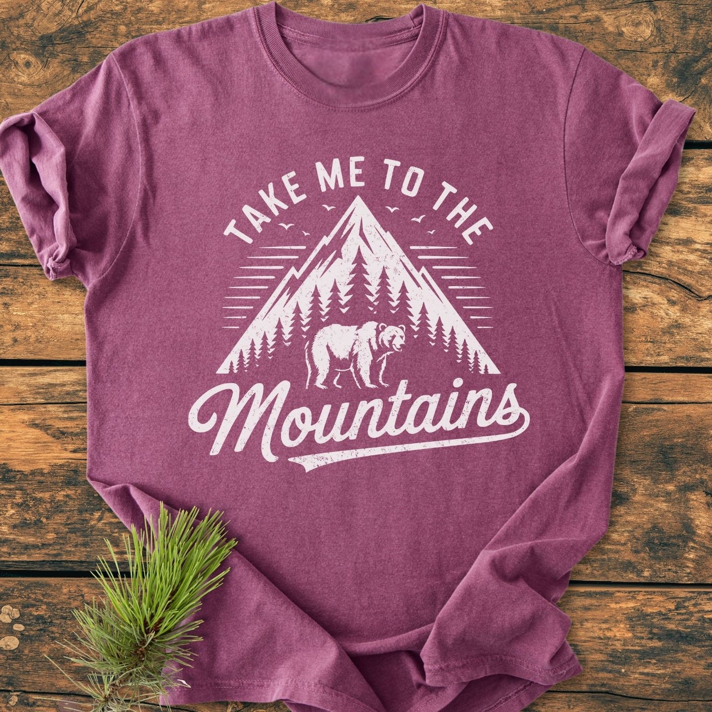 Take Me To The Mountains - Vintage Wash Tee features a mountain and bear design on a maroon/purple shirt, laid flat with pine needles, highlighting its relaxed fit and durable stitching.