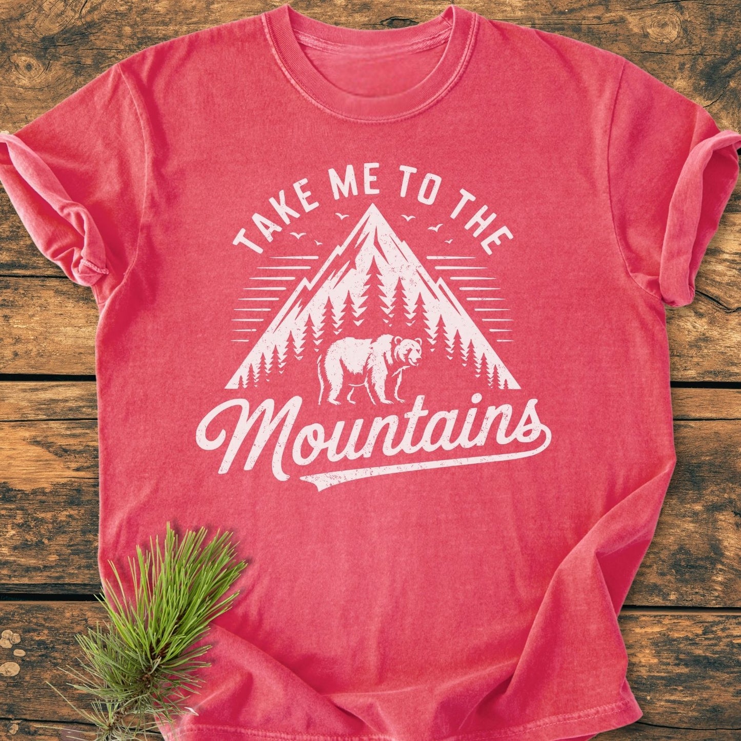 Take Me To The Mountains - Vintage Wash Tee, featuring a mountain and bear graphic, laid flat on wood. Made from soft-washed, garment-dyed cotton, offering a relaxed fit for everyday comfort.