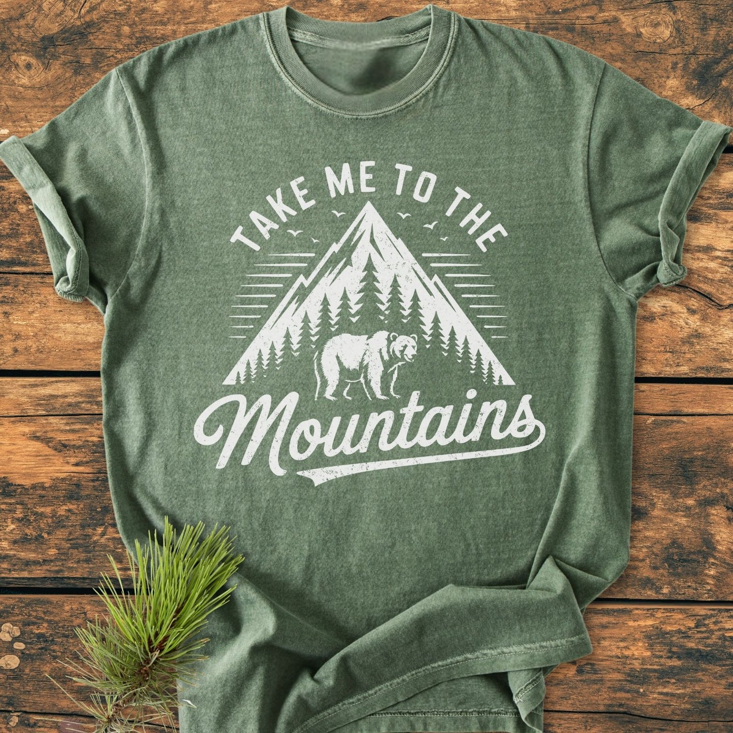 Take Me To The Mountains - Vintage Wash Tee: A heather-green crew-neck T-shirt with a white screen-print of a mountain peak, pine trees, and bear silhouette, laid on rustic wood.