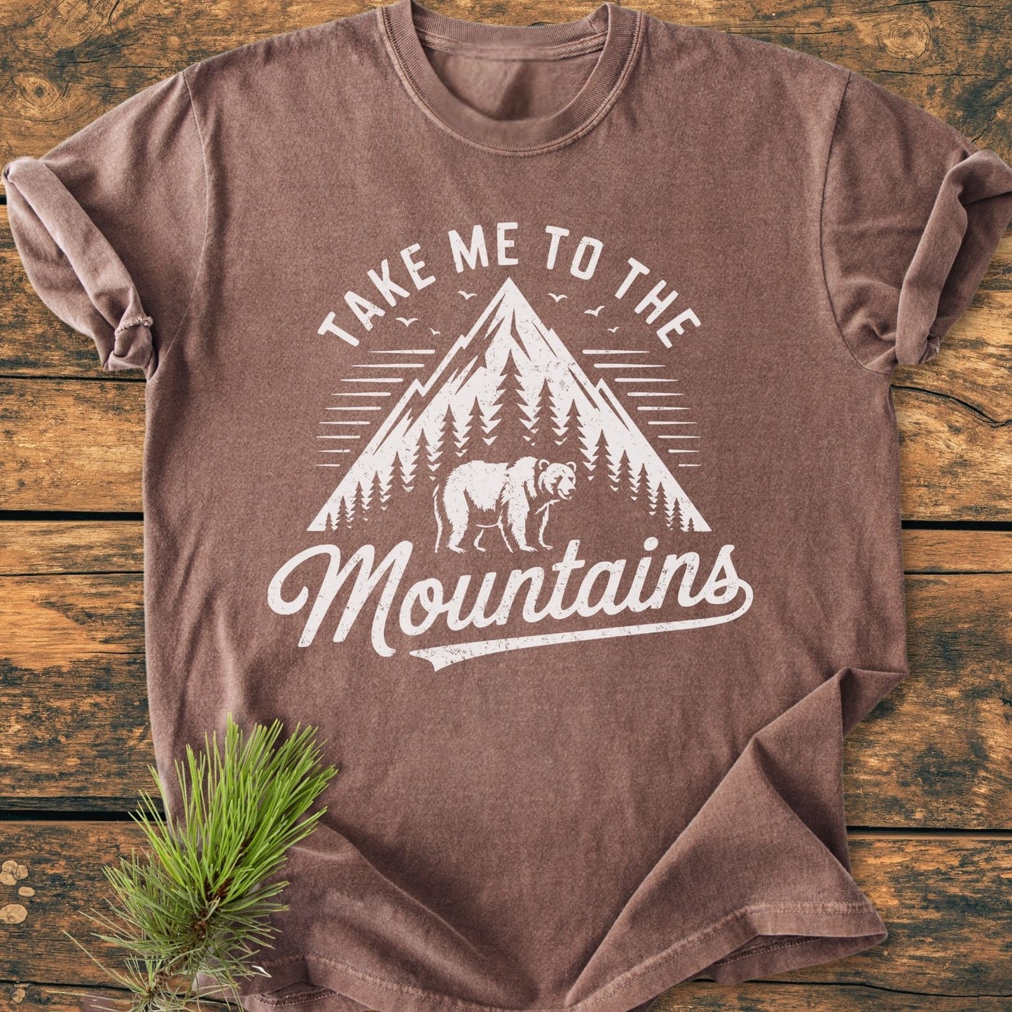 Take Me To The Mountains - Vintage Wash Tee featuring a white mountain peak, pine trees, and bear design, laid on wood with pine sprig beside it, showcasing its relaxed fit and soft fabric.