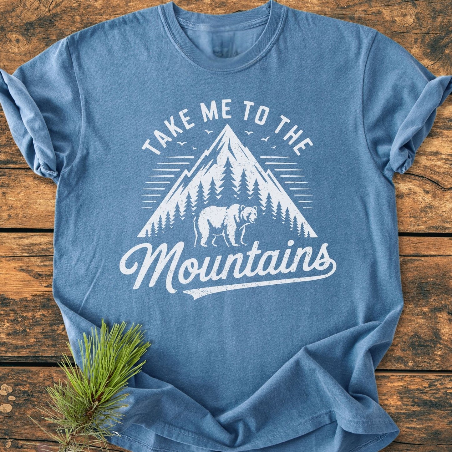 Take Me To The Mountains - Vintage Wash Tee features a stylized mountain and bear graphic on a relaxed, soft-washed garment-dyed shirt, laid flat on a rustic wooden surface.