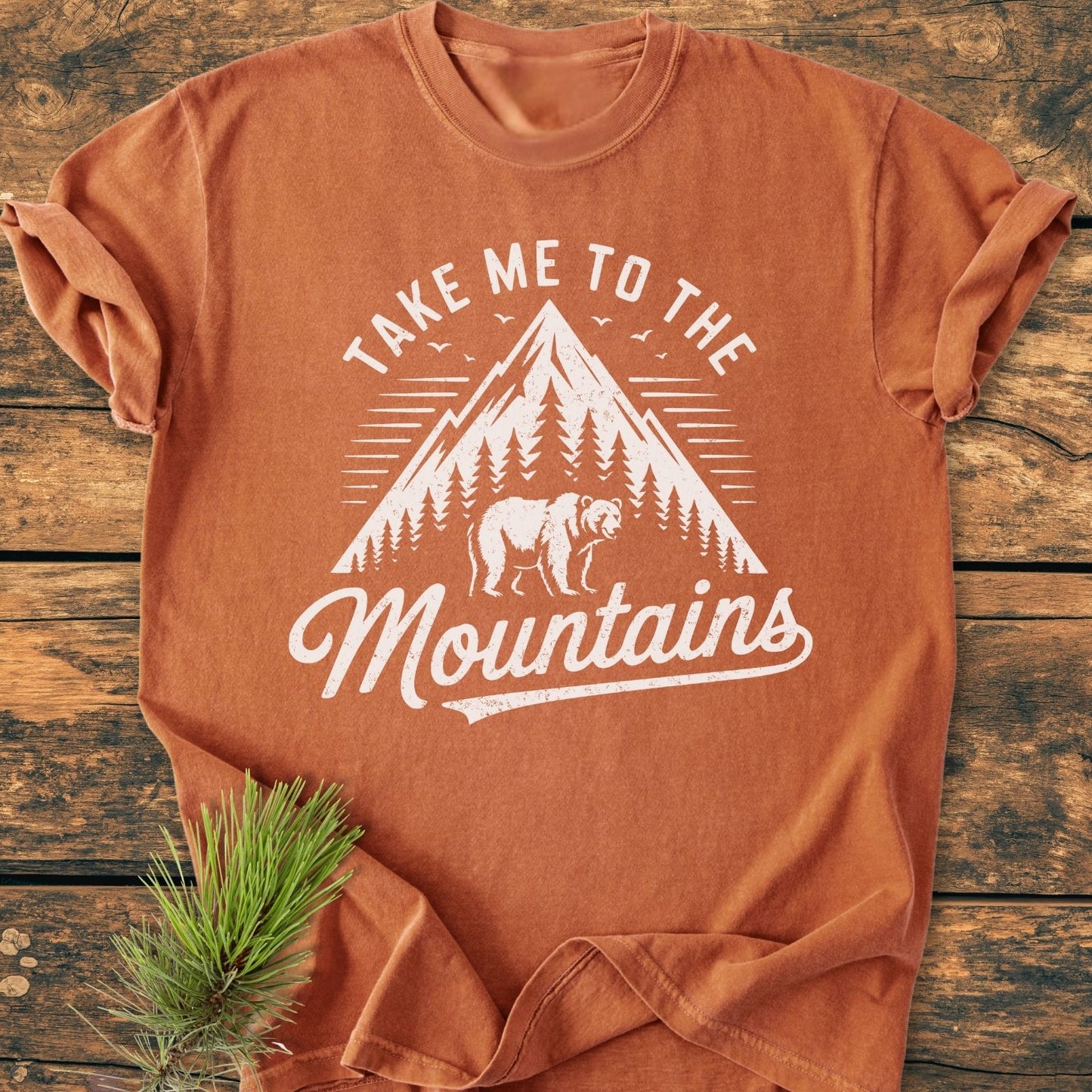 Take Me to the Mountains - Vintage Wash Tee features a stylized mountain scene with pine trees and a bear, laid flat on a wooden surface beside a sprig of pine.