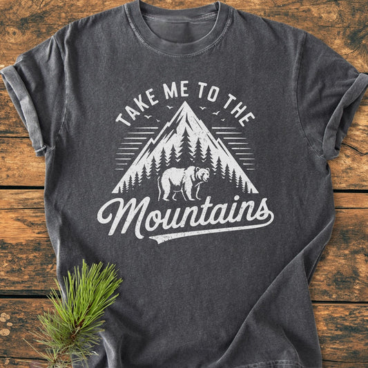 Take Me To The Mountains - Vintage Wash Tee featuring a mountain and bear graphic on soft, garment-dyed fabric, laid out on wood-grain; accented with a pine sprig for rustic appeal.