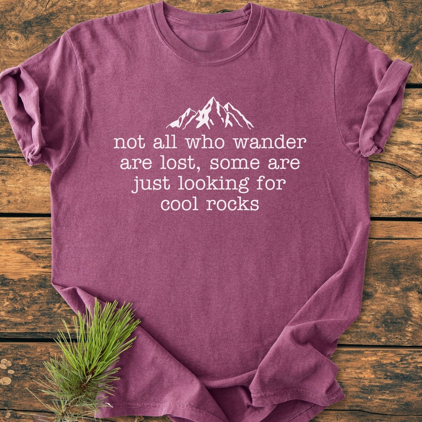 Looking For Cool Rocks Vintage Wash Tee featuring a white mountain graphic and text on a heather-purple fabric, laid on wood with a pine sprig accentuating its outdoorsy theme.