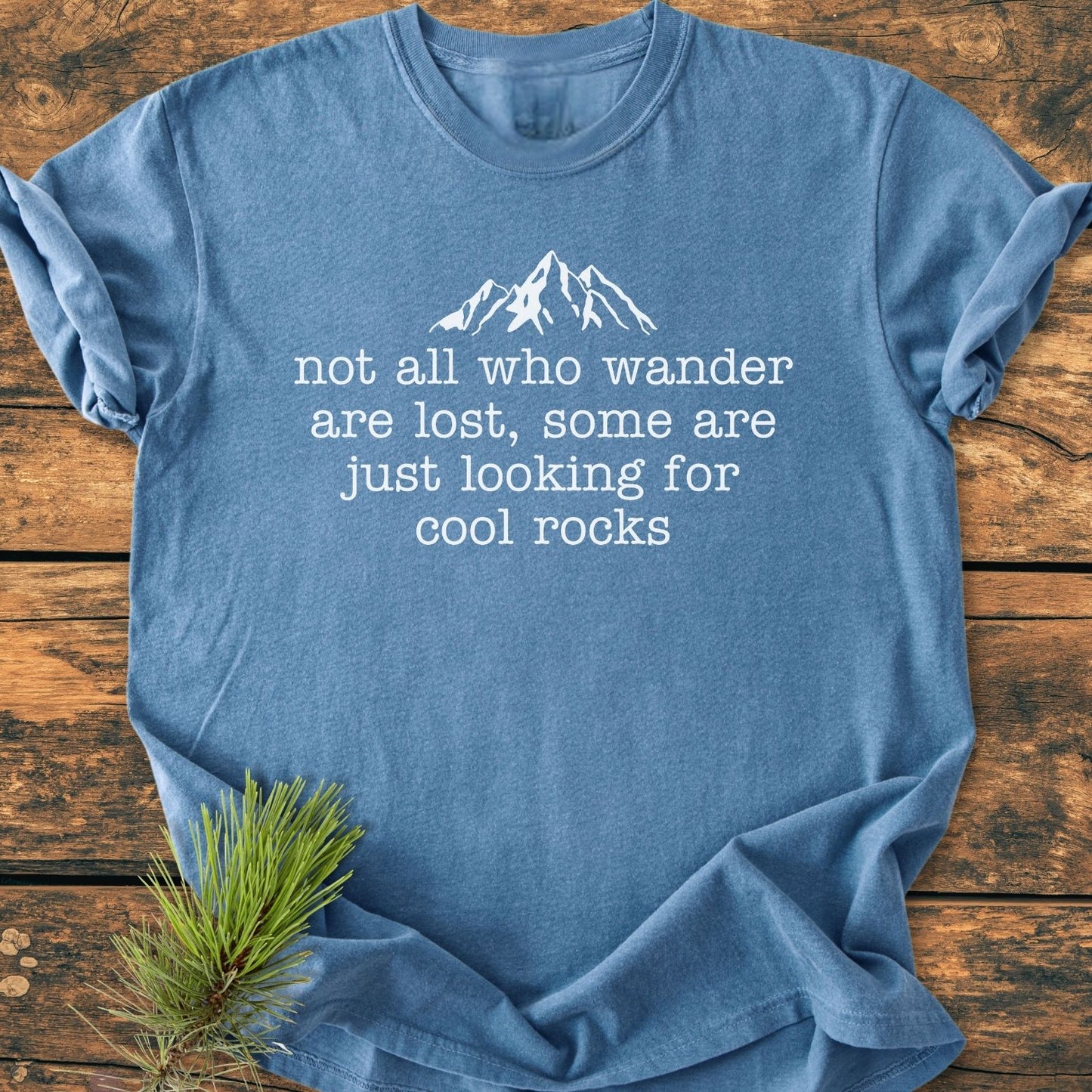Looking For Cool Rocks Vintage Wash Tee features a heather-blue color with a mountain graphic and quote, presented on a wooden surface with a pine sprig for an outdoorsy touch.