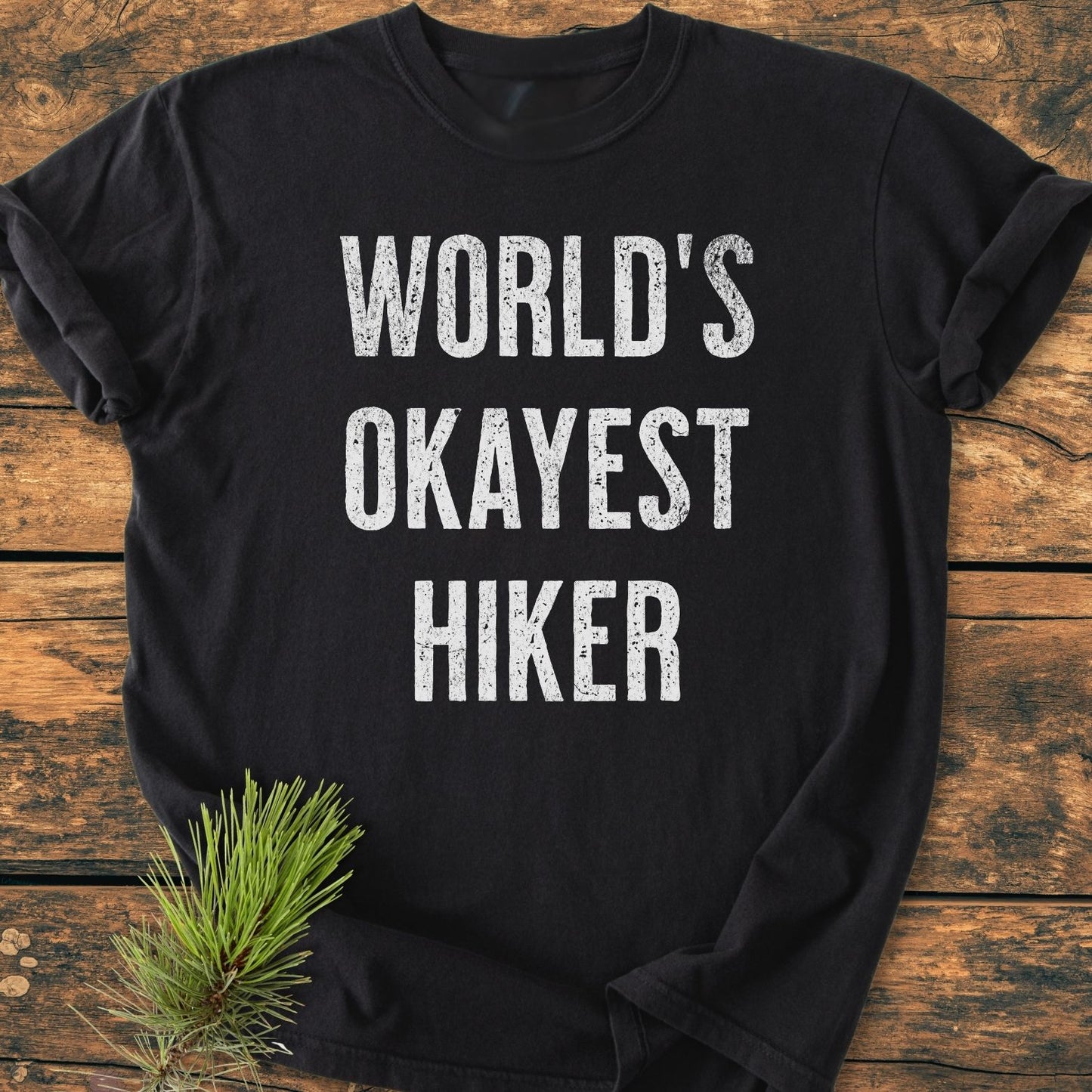 Black World's Okayest Hiker Vintage Wash Tee displayed on wood with a pine branch. Features bold white slogan, relaxed fit, durable double-needle stitching. Made from soft, pre-shrunk 100% ring-spun cotton.