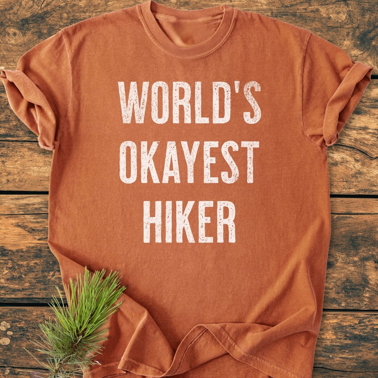 World's Okayest Hiker Vintage Wash Tee displayed on wooden planks, featuring distressed white lettering. A pine sprig accents the soft, relaxed-fit shirt, showcasing its rustic charm.