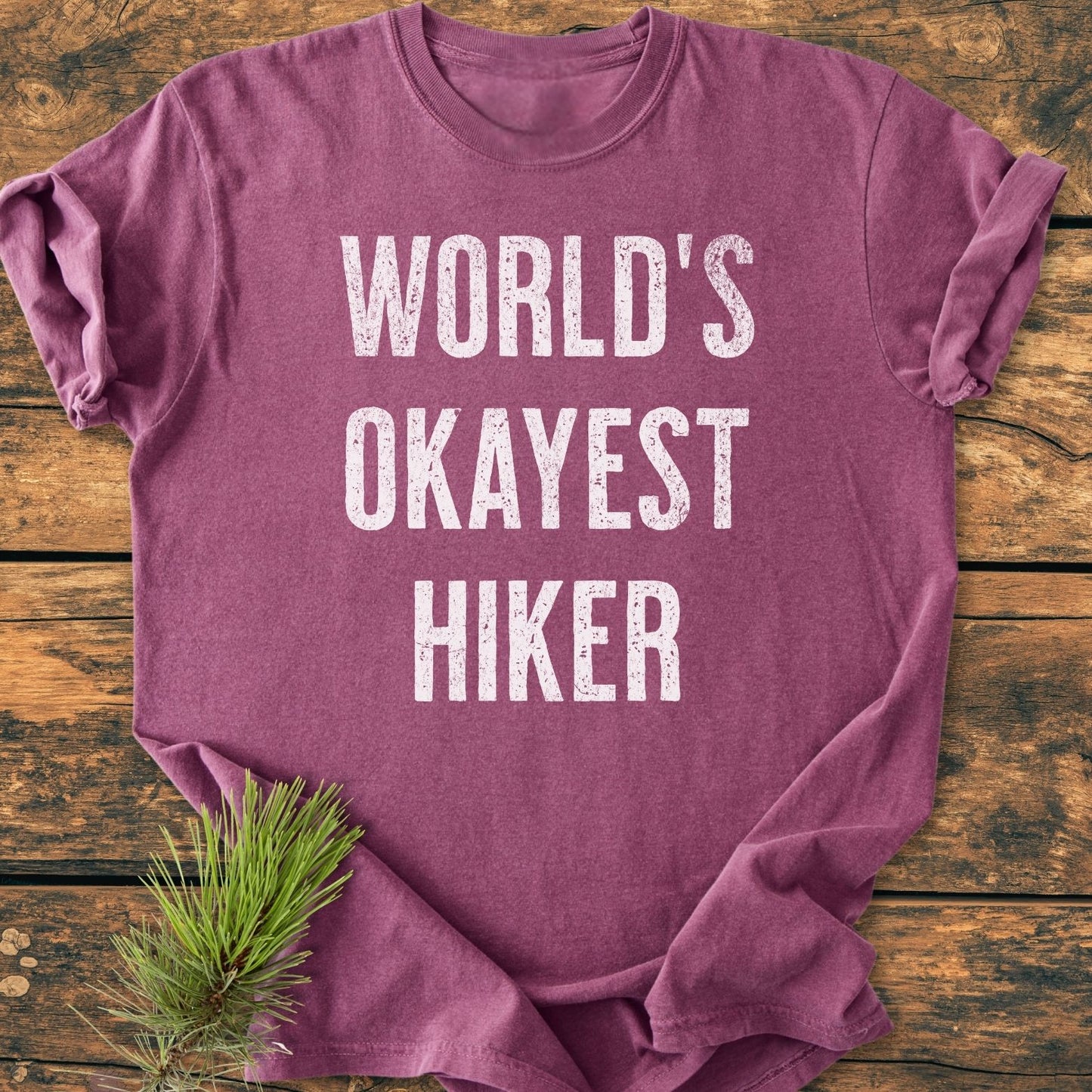 Heather-purple World's Okayest Hiker Vintage Wash Tee folded on wooden surface, featuring distressed white lettering. A pine sprig accents the bottom left, highlighting the tee's rustic charm and relaxed fit.
