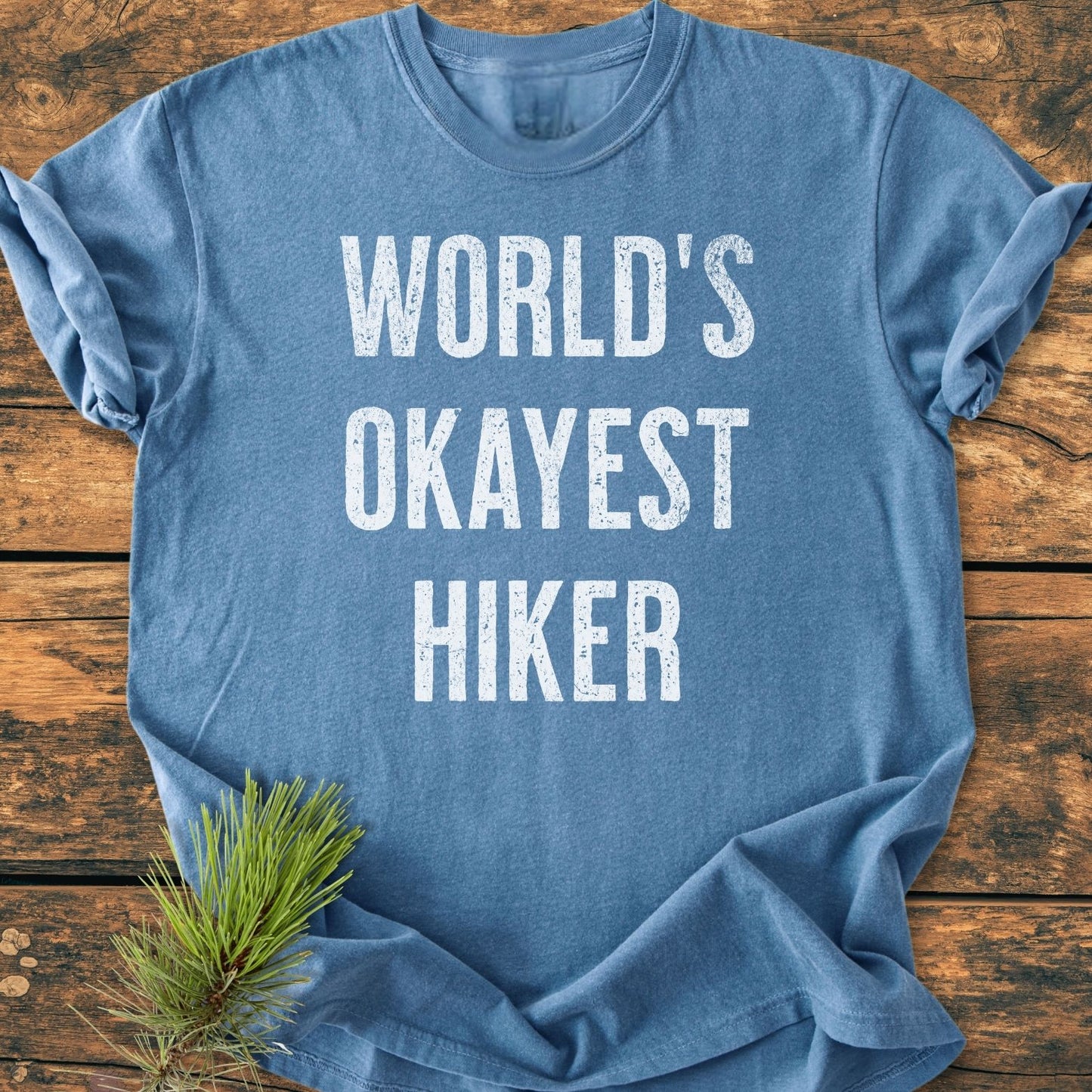 Heather-blue World's Okayest Hiker Vintage Wash Tee with distressed white lettering, laid on rustic wood. Features soft, garment-dyed fabric, offering comfort and relaxed fit. Pine sprig accents the lower corner.