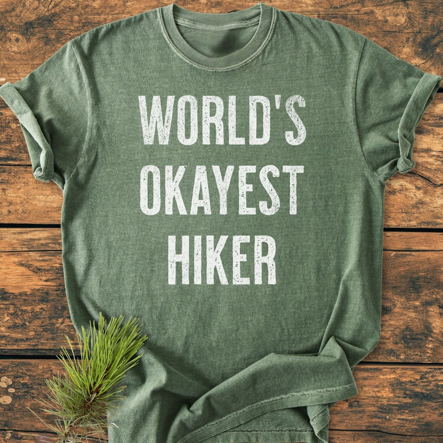 World's Okayest Hiker Vintage Wash Tee in heather-green, screen-printed with a distressed slogan, laid on wood; features durable, soft-washed cotton, relaxed fit, complemented by a pine sprig for a rustic touch.