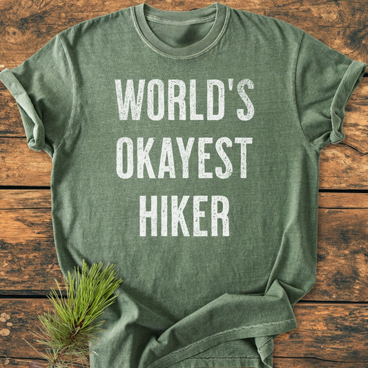 World's Okayest Hiker Vintage Wash Tee in heather-green, screen-printed with a distressed slogan, laid on wood; features durable, soft-washed cotton, relaxed fit, complemented by a pine sprig for a rustic touch.